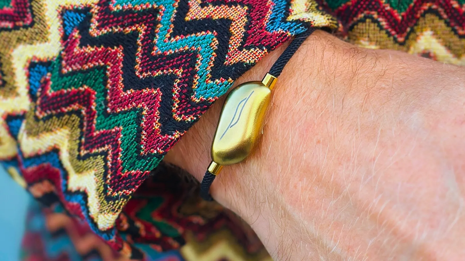 The Sou Band worn on a wrist, its brushed gold pebble on a dark cord, beside a brightly patterned zigzag sleeve.