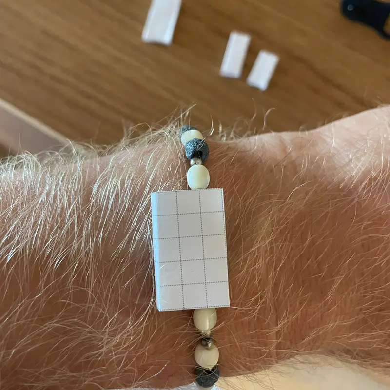 A white rectangular prototype module taped to a wrist beside a beaded bracelet.