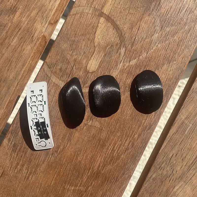 Three matte black pebble samples and a bare circuit board on a wooden table, laid against a tape measure.