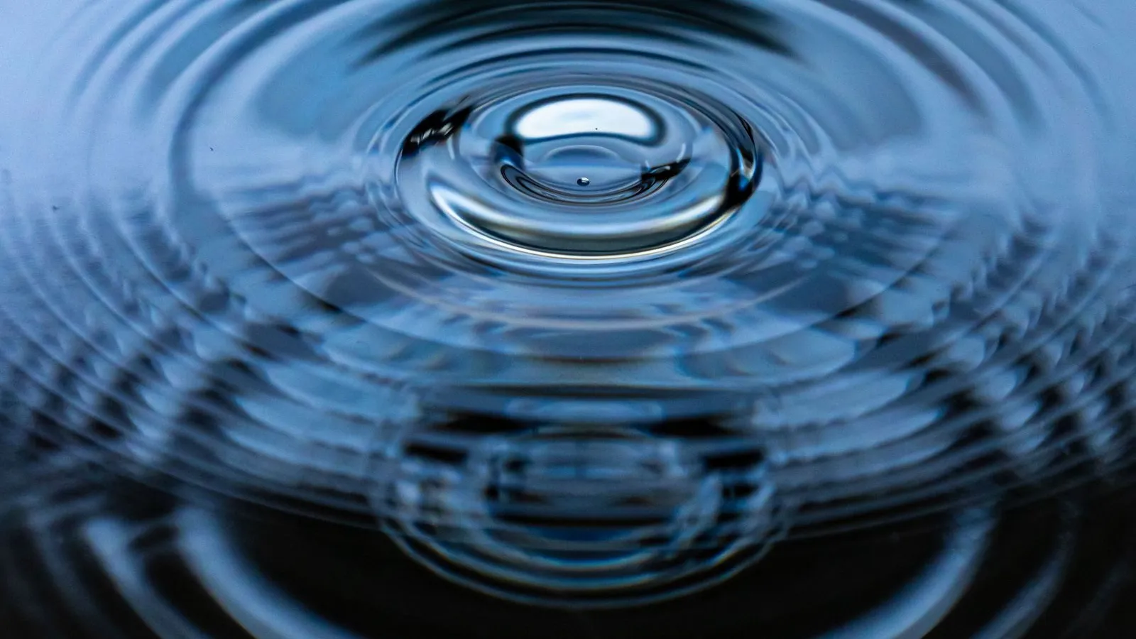 A single water droplet meeting still water, sending concentric ripples outward across a calm blue surface.