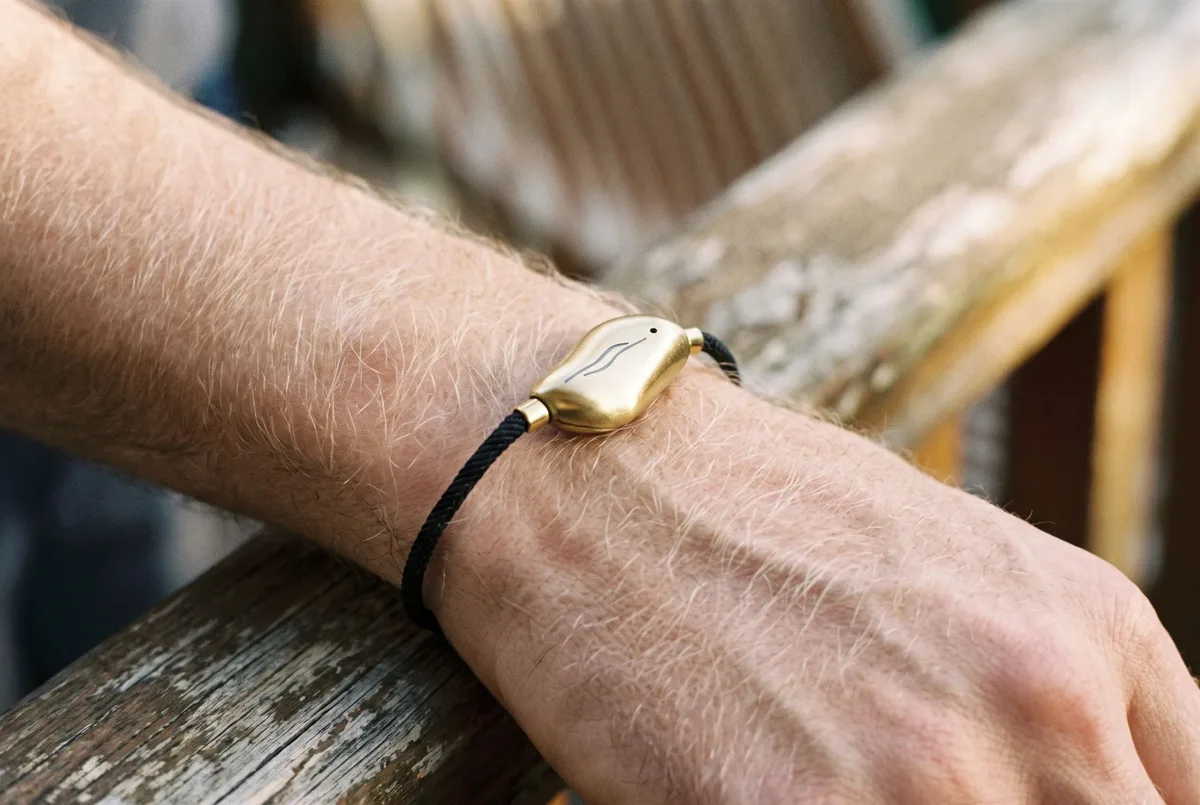 A Sou Band worn on a man's wrist as his hand rests on a wooden bannister: a small gold pebble on a thin black cord.