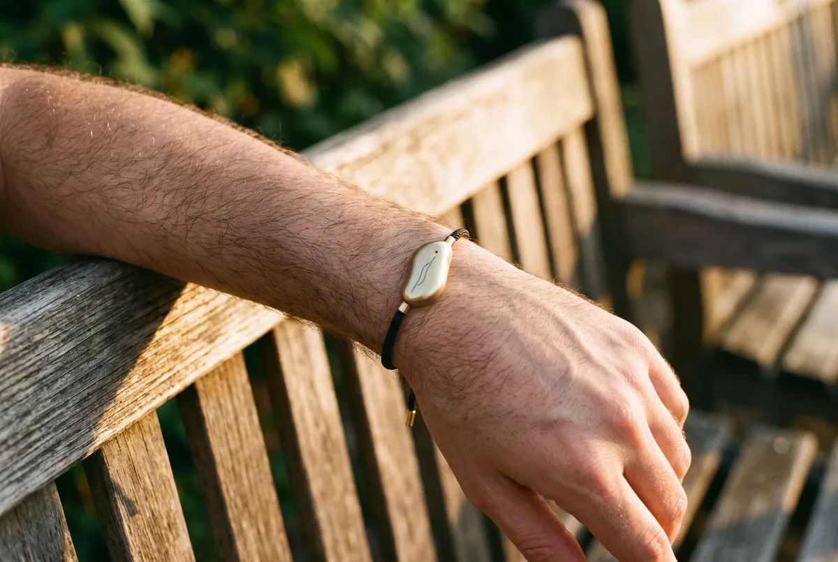A Sou Band worn on a man's wrist as he sits on a park bench with his arm along the back of it: a small gold pebble on a thin black cord.