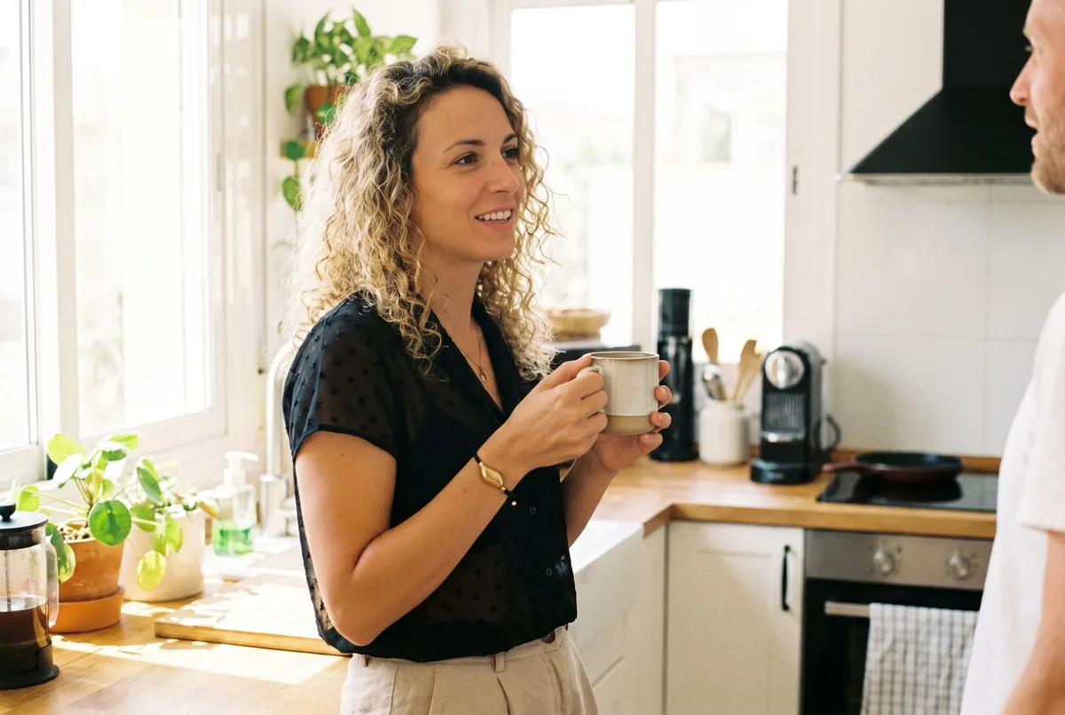 A Sou Band worn on a woman's wrist as she stands talking in a bright kitchen with a mug in her hand.