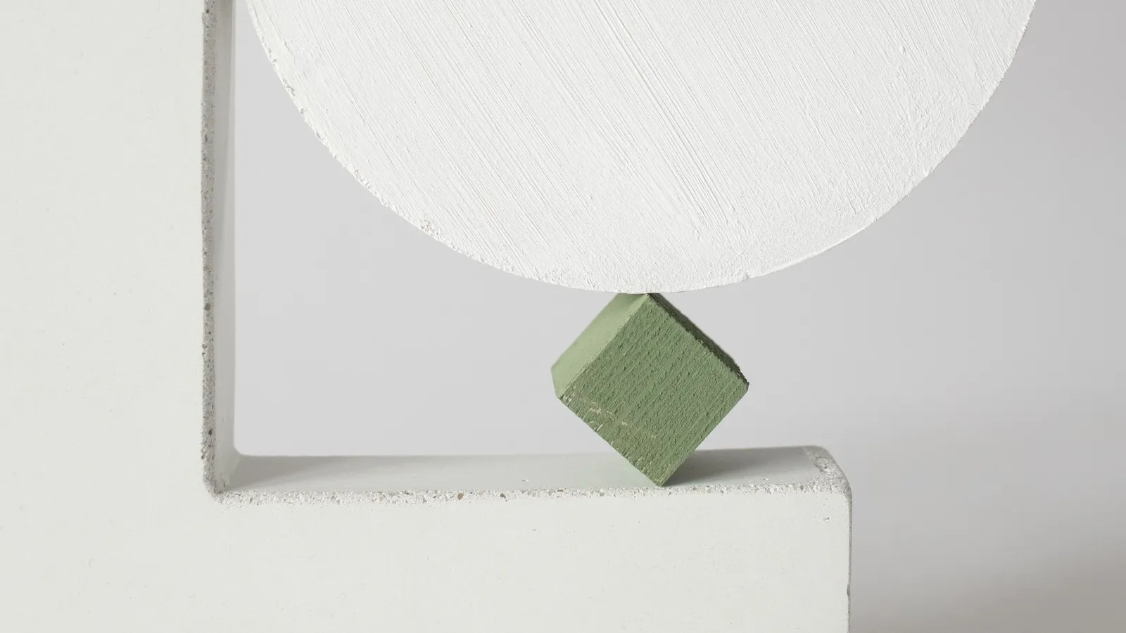 A large white painted disc balanced on a single small green wooden cube tipped onto its corner, resting on a white plaster ledge.