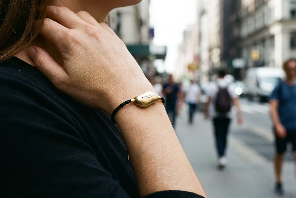A Sou Band on a woman's wrist as she raises her hand to her collarbone on a city street: a gold pebble on a thin black cord.