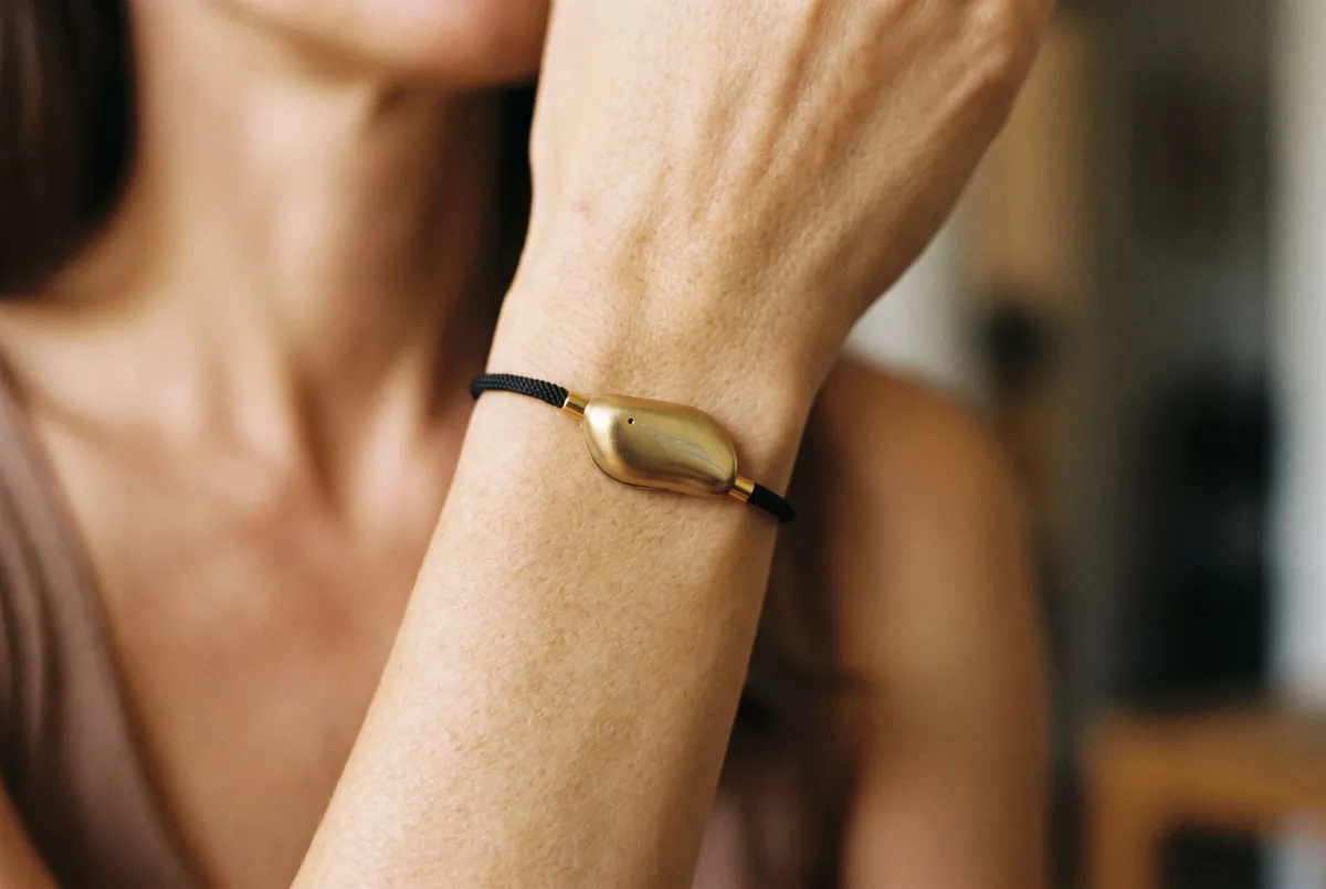 A Sou Band worn on a woman's wrist as she raises her hand to her collarbone: a small gold pebble on a thin black cord.