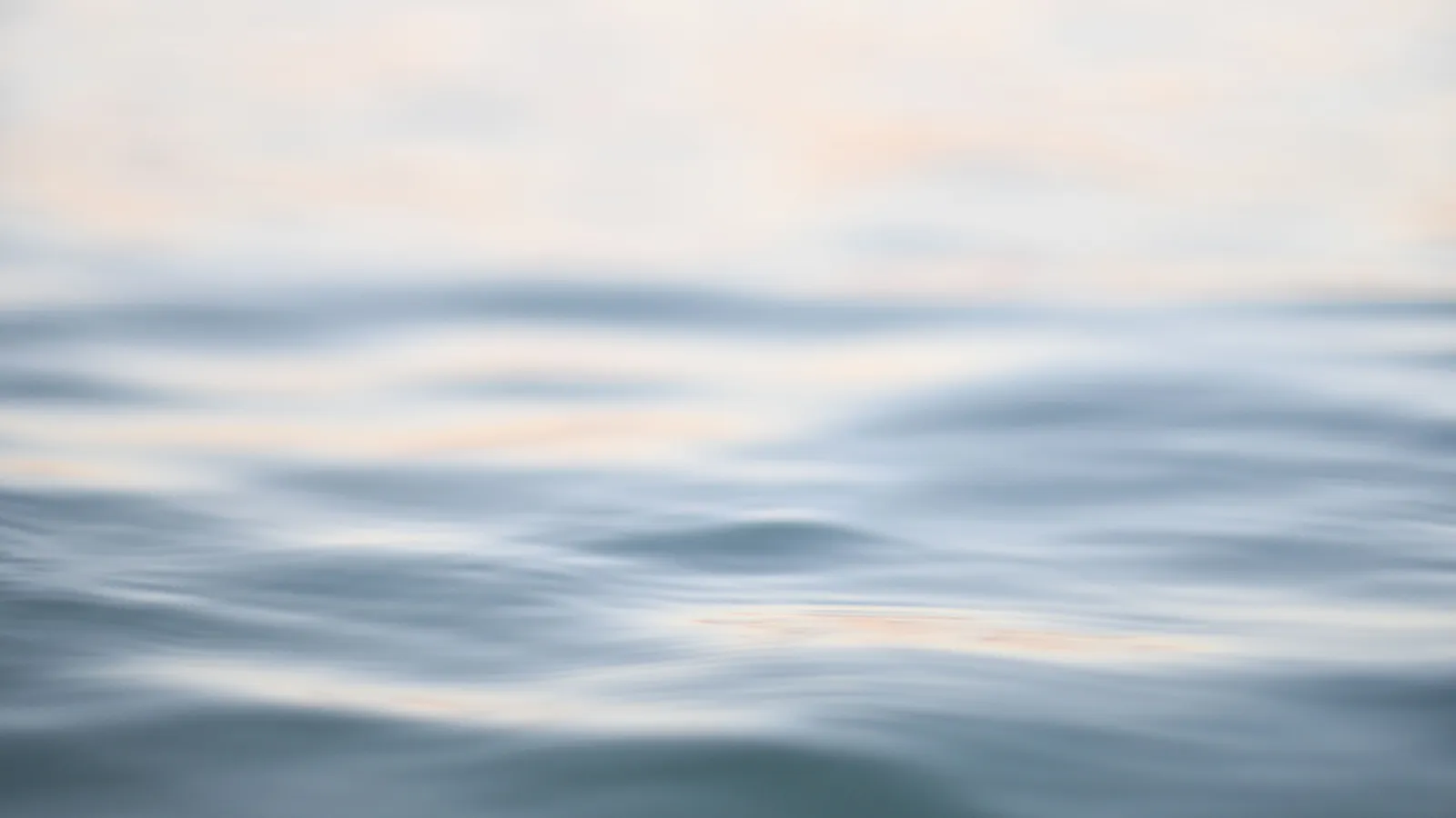 Soft-focus water at dawn, pale blue swells lit by warm cream and peach light.